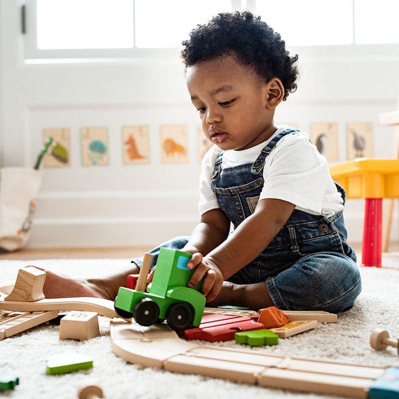 A toddler in dungarees kneeling on a rug, pushing a wooden train around a track