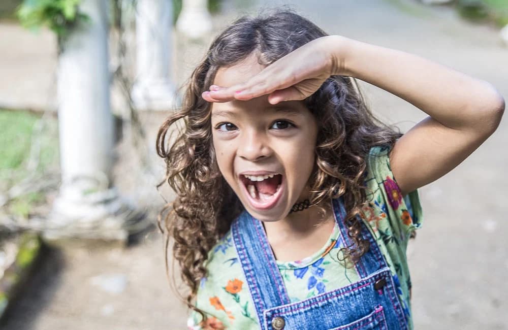 A smiling girl in denim dungarees shading her eyes with one hand while playing outdoors