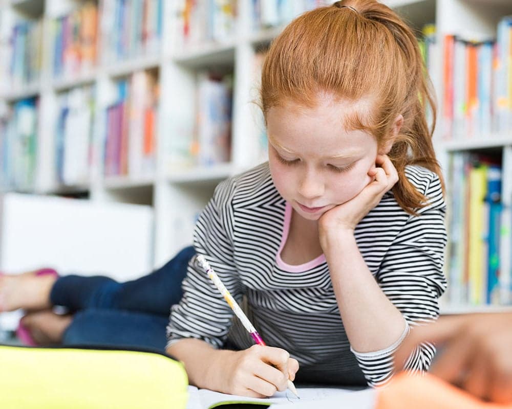 A girl lying on the floor writing in a notebook, with shelves of books behind her