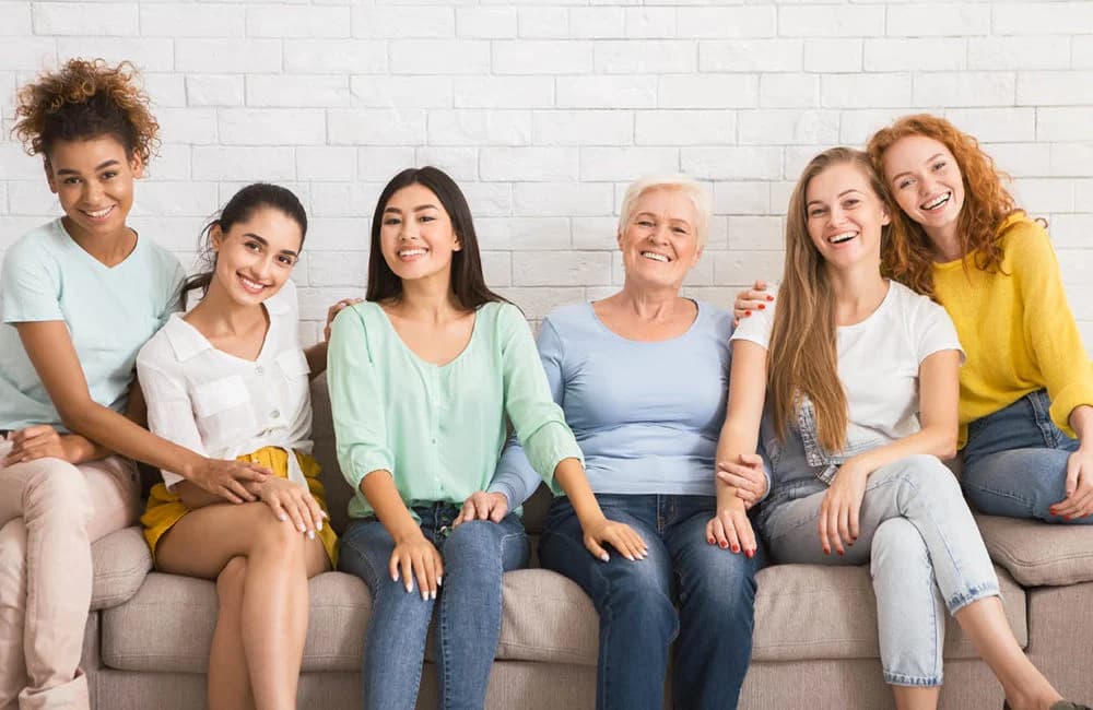 Six women of different ages sitting close together on a sofa, laughing