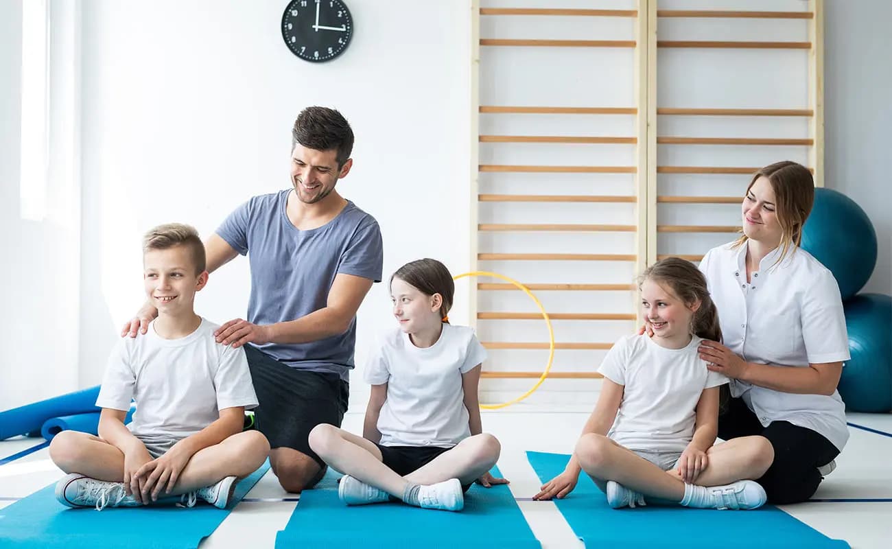 Children sitting on gym mats in a hall with two adults