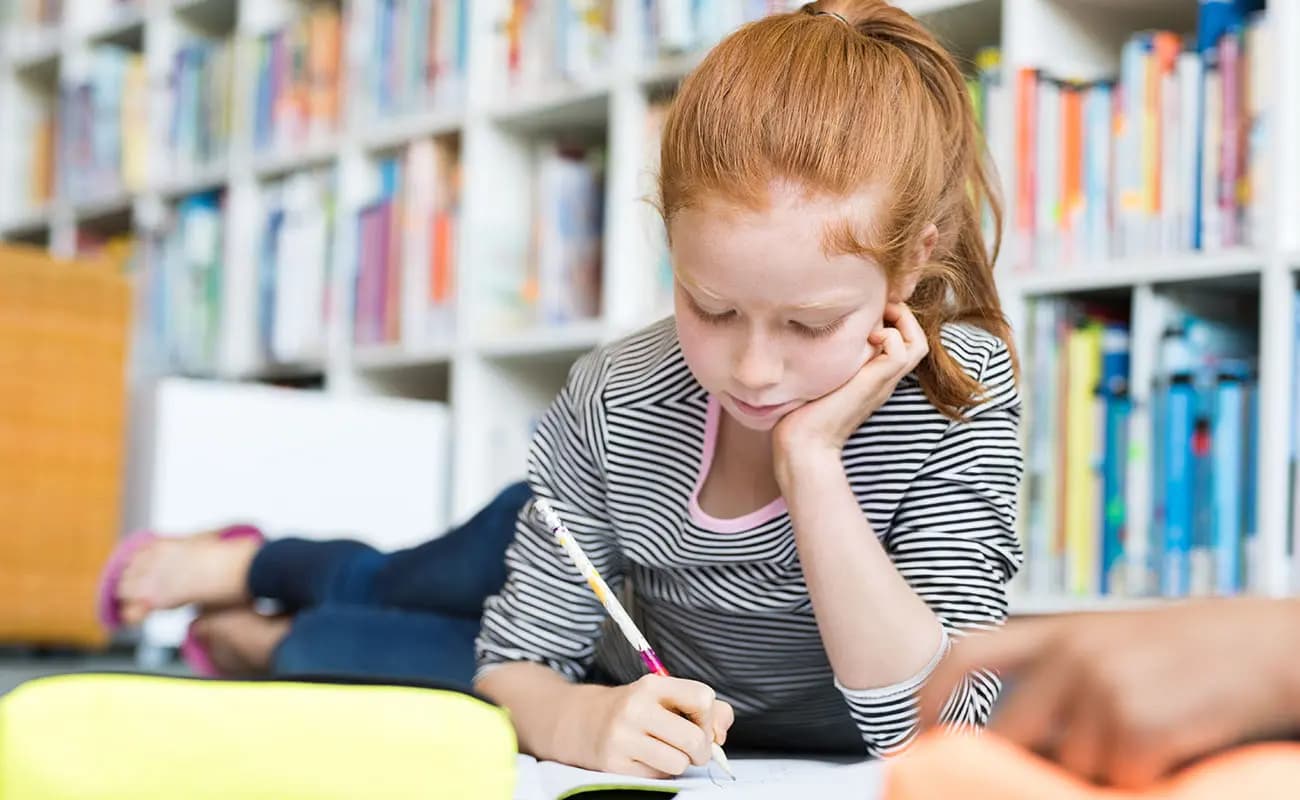 A child writing in a notebook beside shelves of books