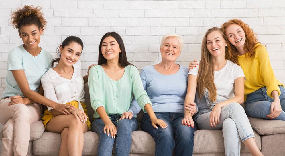 Six women of different ages sitting together on a sofa, smiling at the camera